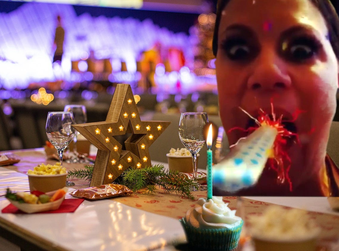 Silly photo of me blowing a paper party favor noise maker, those things that unwind from your mouth, out to a candle in a birthday cupcake, all set atop a lavishly decorated birhtday table with city lights in the background.