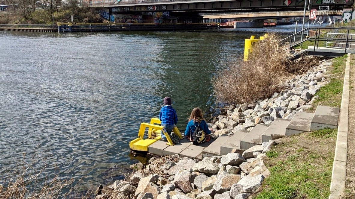Two children sitting by a river near to two bridges.