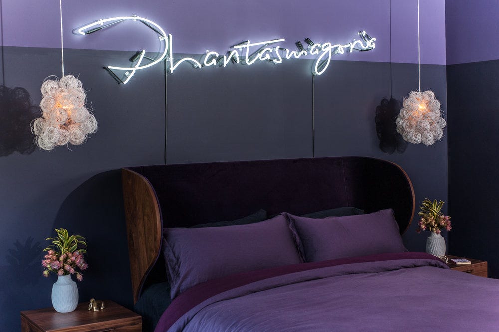 Close up of the bed with a custom, curved wooden headboard, sculptural light fixtures, the neon sign, and two toned plum wall colors.
