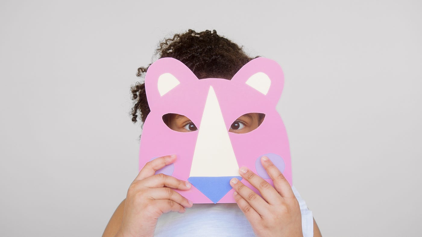 A girl hides behind a paper mask of a pink lion.
