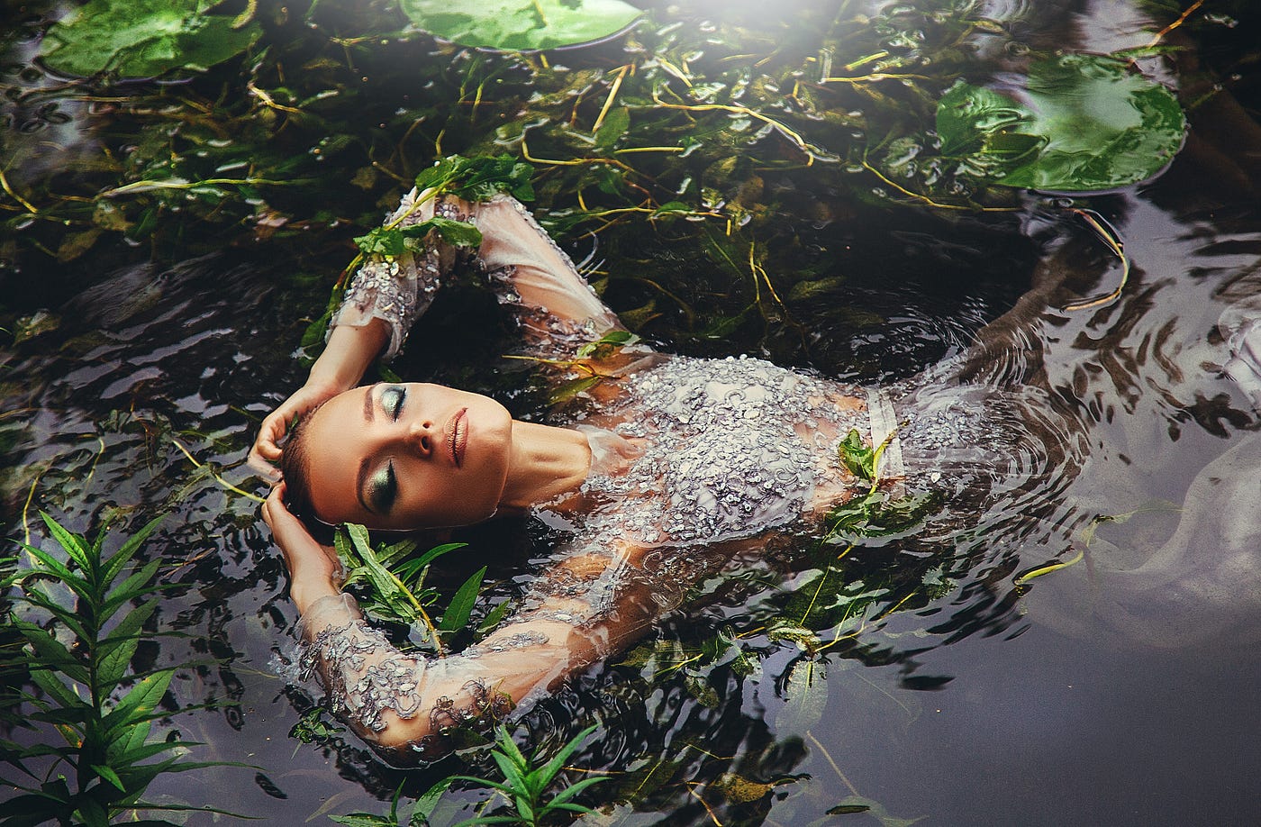 Beautiful woman is immersed in water on her back in seaweed. She is face up, eyes closed. Beautiful makeup, elbows bent, hands resting on the top of her head. Magic, beauty, fantasy aesthetic. Captivating.