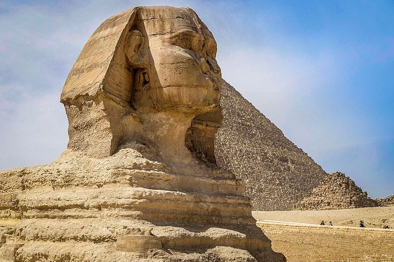 The Great Sphinx at Giza. An ancient, huge stone statue with a head resembling that of a man, with a serpent’s flares, on top of a lion’s body. The Great Pyramid is in the background.