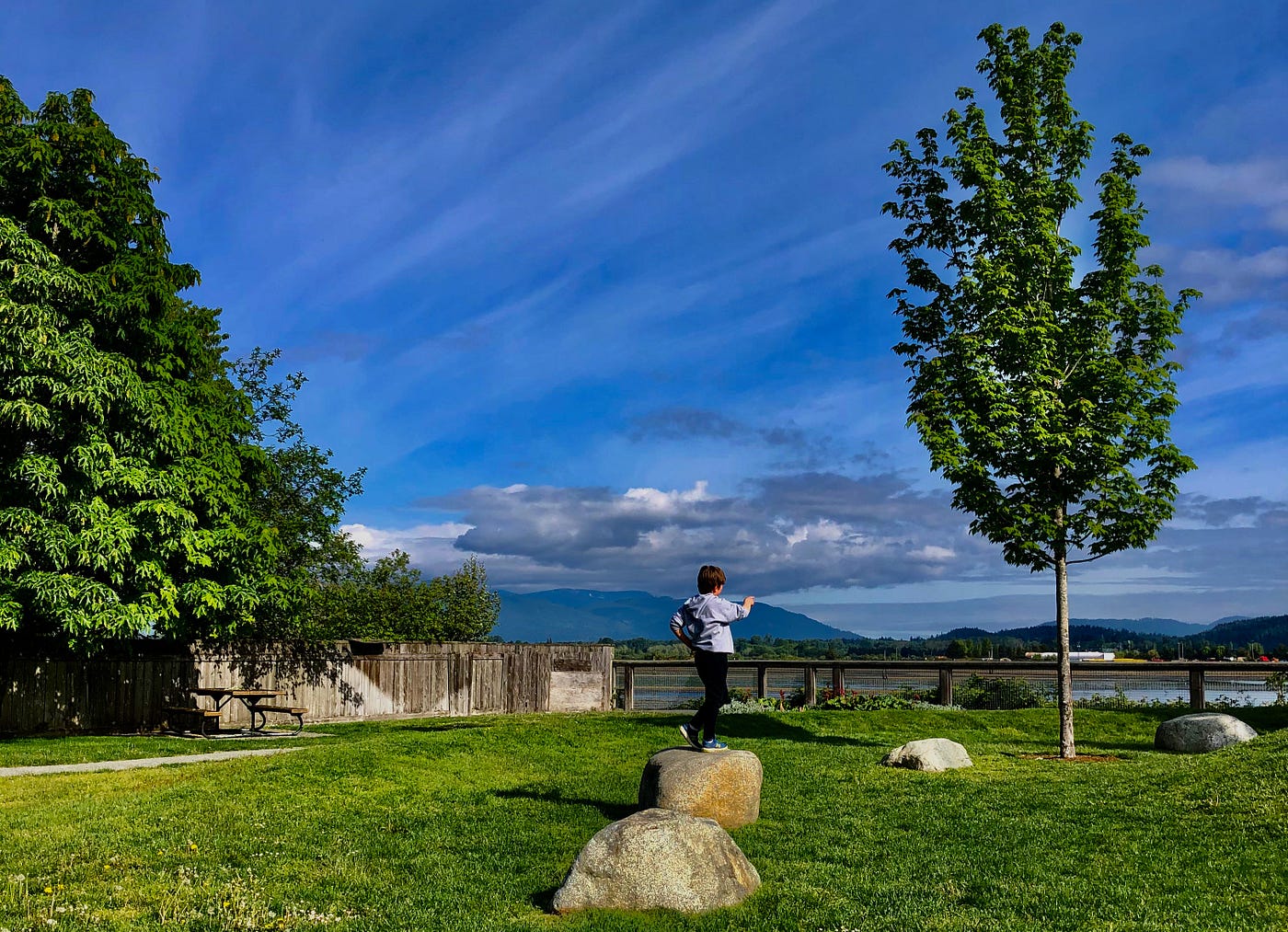 a boy stands on a rock in a park. a blue sky is overhead and trees nearby