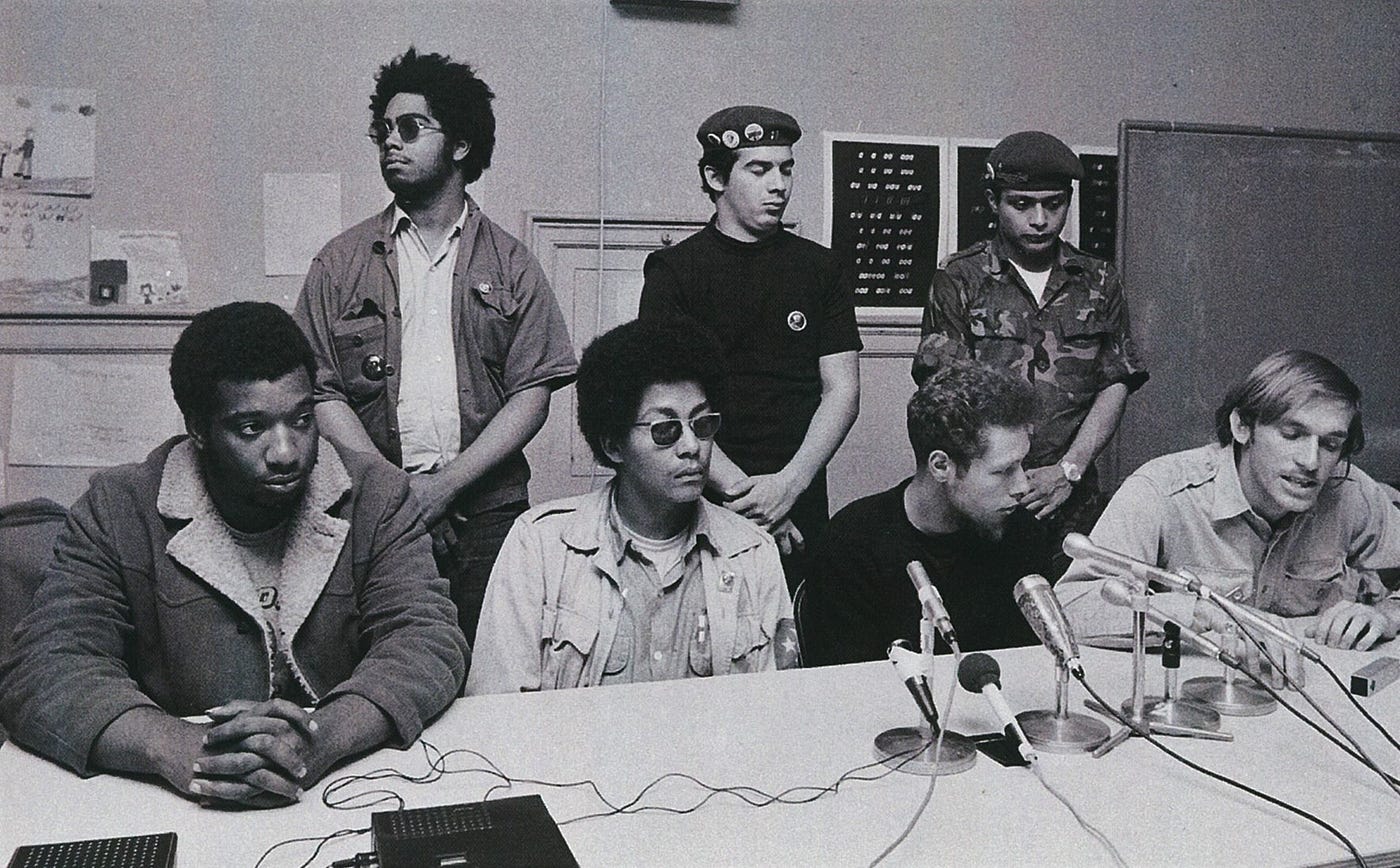 Seven members of Chicago’s Rainbow Coalition sits behind a table with microphones at what looks like a press event.