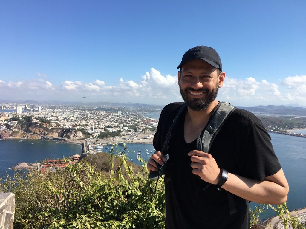 Omar, a Latino man with short black hair and a long beard, is outside on a bright day on the top of a hill, with the sea below. He wears a black cap and smiles at the camera. He also wears a black t-shirt and a watch on his left wrist.