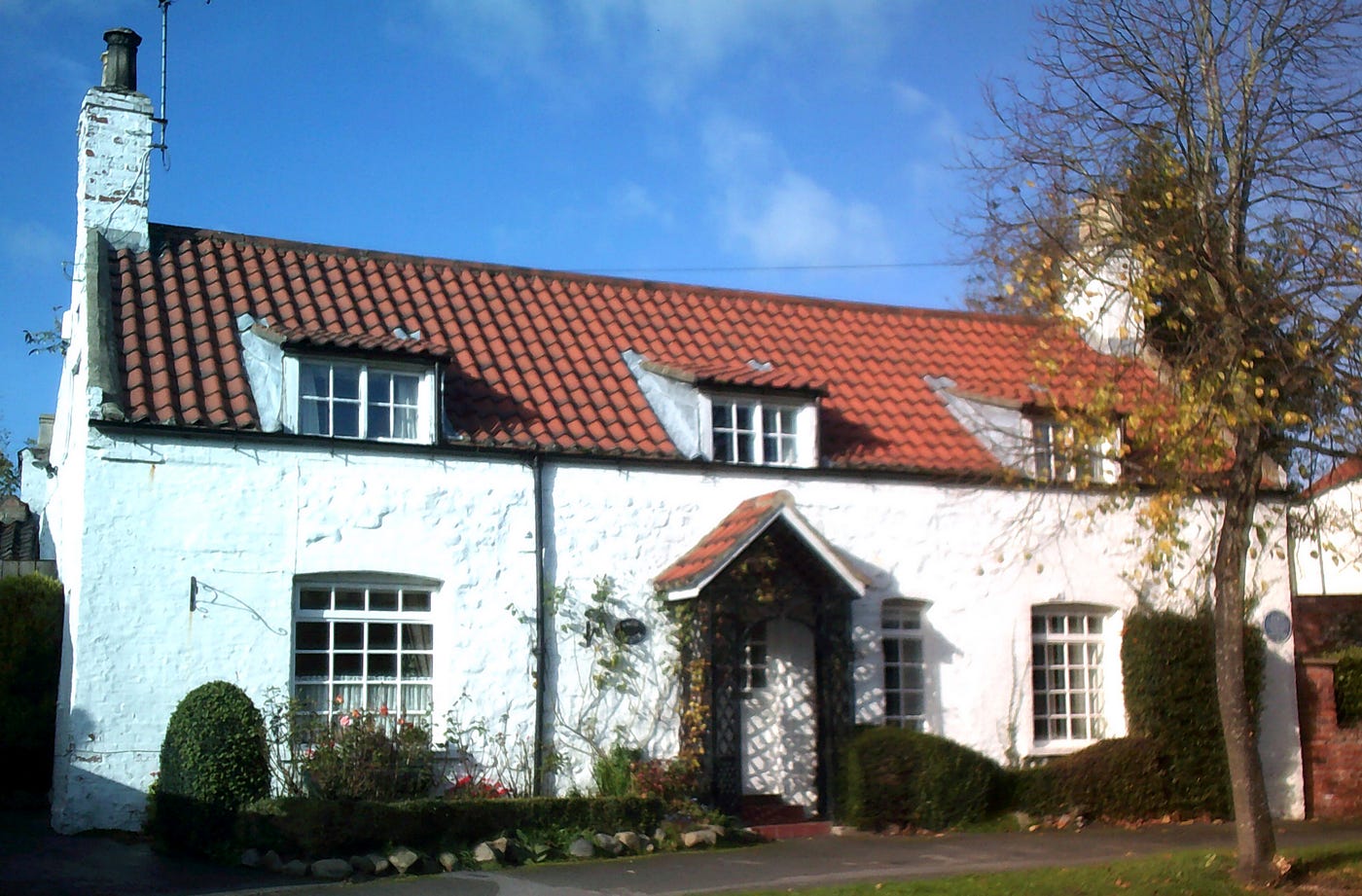 Photograph of a small detached English cottage called ‘White Cottage’ by the side of the road with blue sky in the background