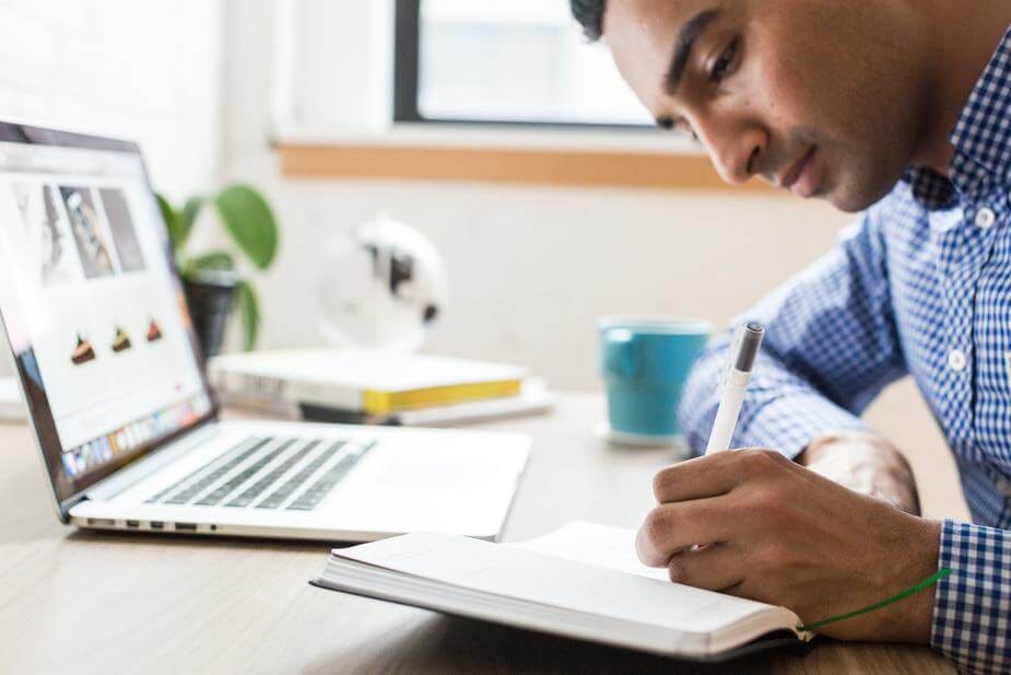 A man writing in a notebook with a pen. He has an open laptop on the desk in front of him.