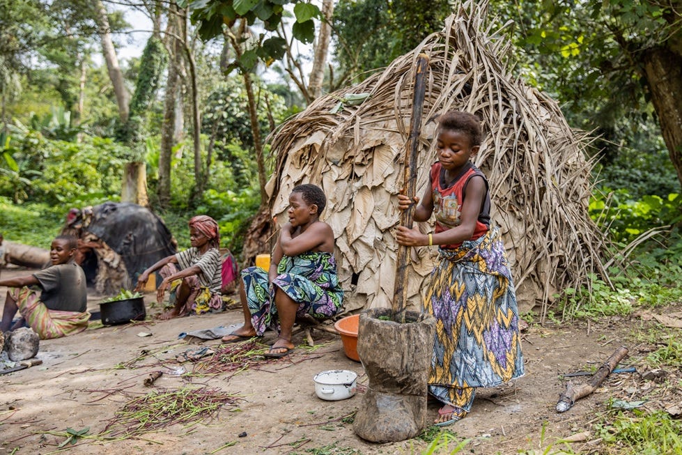 An Indigenous girl prepares a meal as her family sits outside their thatched home.