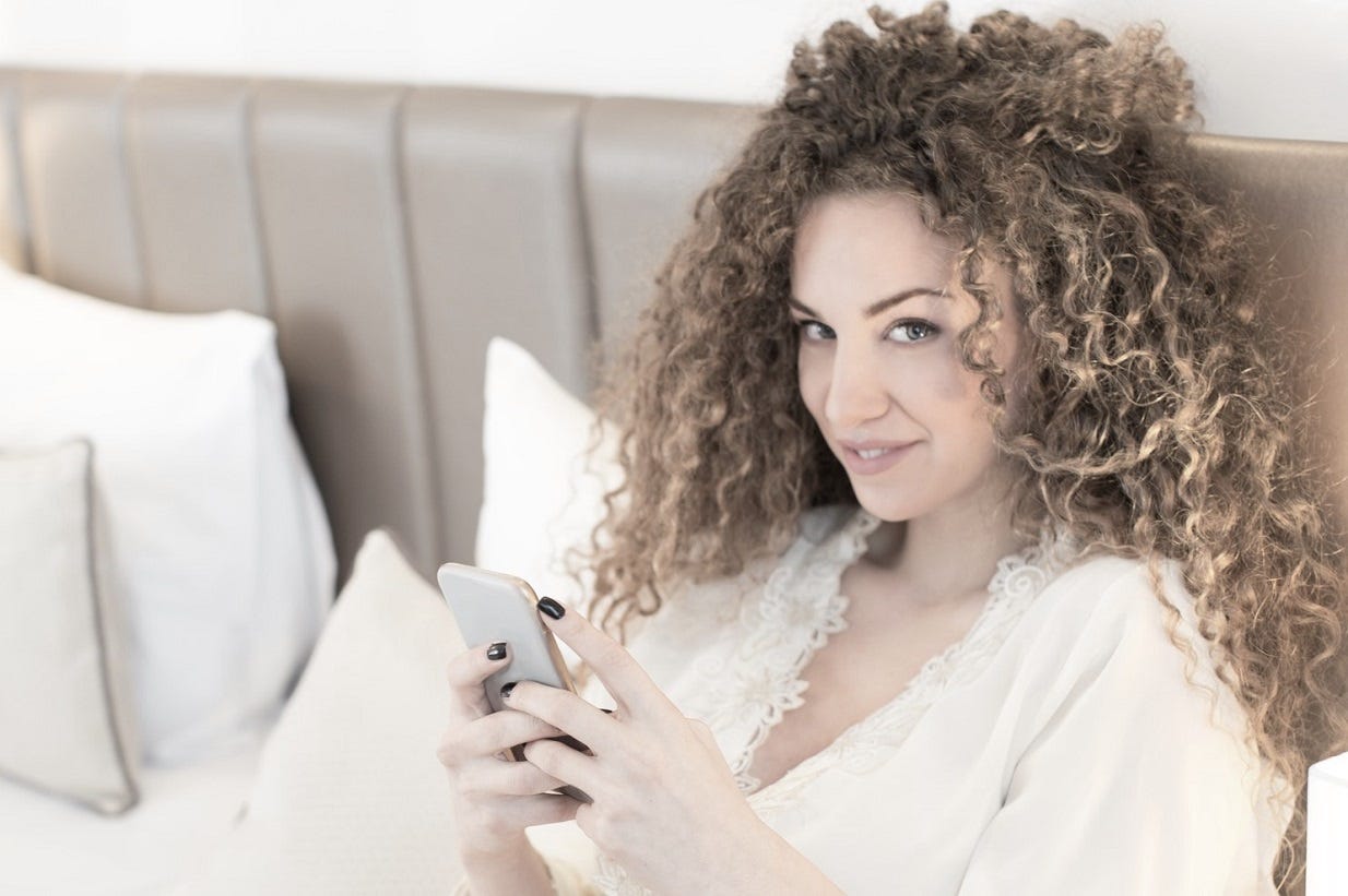Young woman with curly hair taking a selfie photo in the bedroom.
