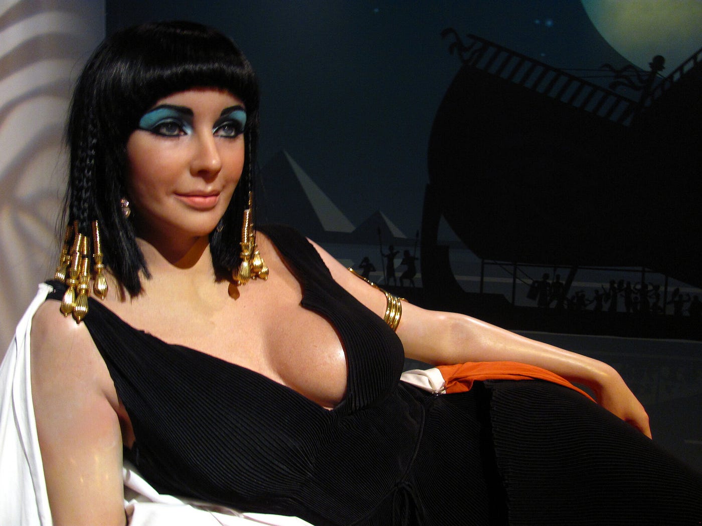 A lifelike wax effigy of Elizabeth Taylor as Cleopatra. She sports a sexy low-cut black gown; sexy, dramatic turquoise eyeshadow; and gold bobbles at the end of shoulder-length black braids.