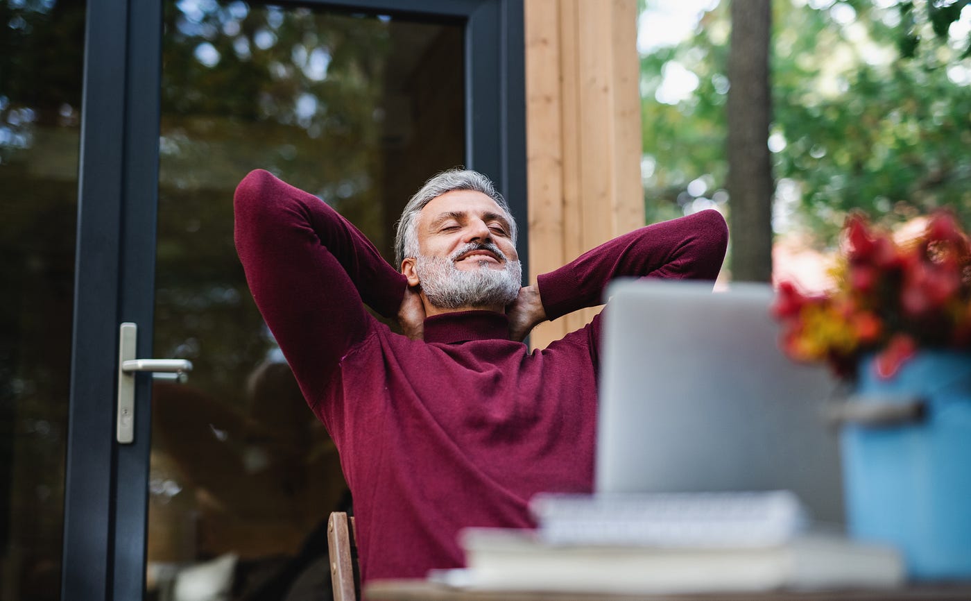 Man with greying beard relaxing outside, hands behind his head, with a relaxed expression.