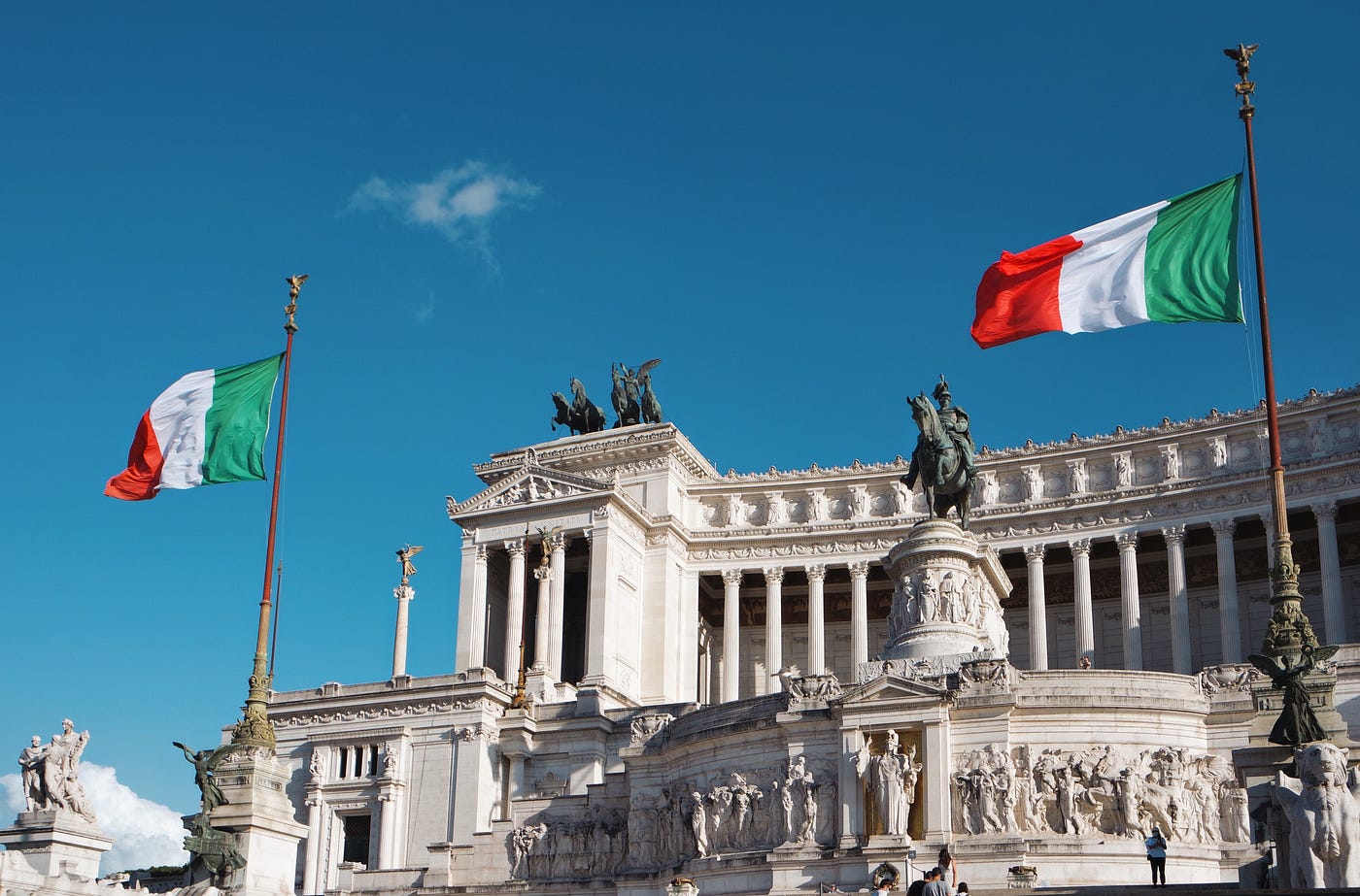 This photo shows the famous Vittorio Emmanuele II Monument in the heart of Rome. There are two Italian flags on each side of the monument and a figure on a horse at the center. There are Roman columns at the front of the building and classical Roman architecture along with embedded bas relief on the building.