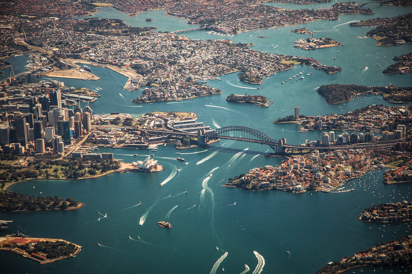 An aerial view of the city of Sydney Australia. In the photo you can see Darling Harbor, the Harbor Bridge, and the Sydney Opera House. There are many boats in the harbor leaving white ripples in the water.