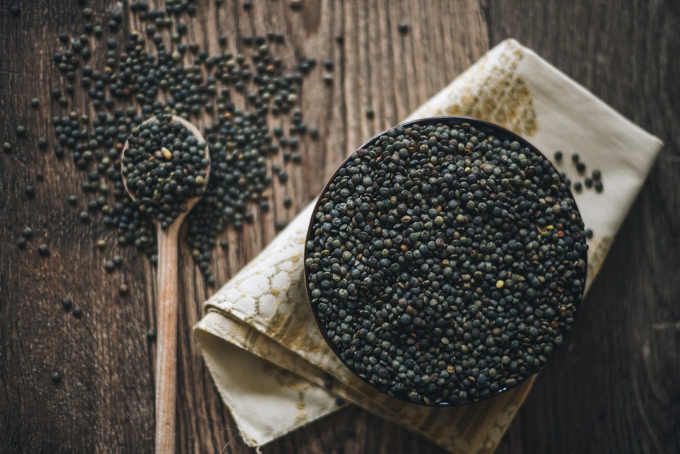 French lentils in a bowl on a cloth napkin, next to a wooden spoon heaped with lentils.