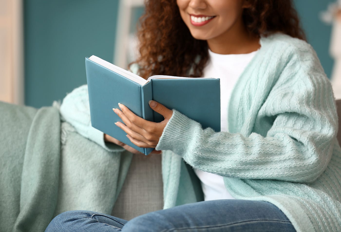 Young African-American woman in blue cardigan reading book on sofa at home. By Pixel-Shot