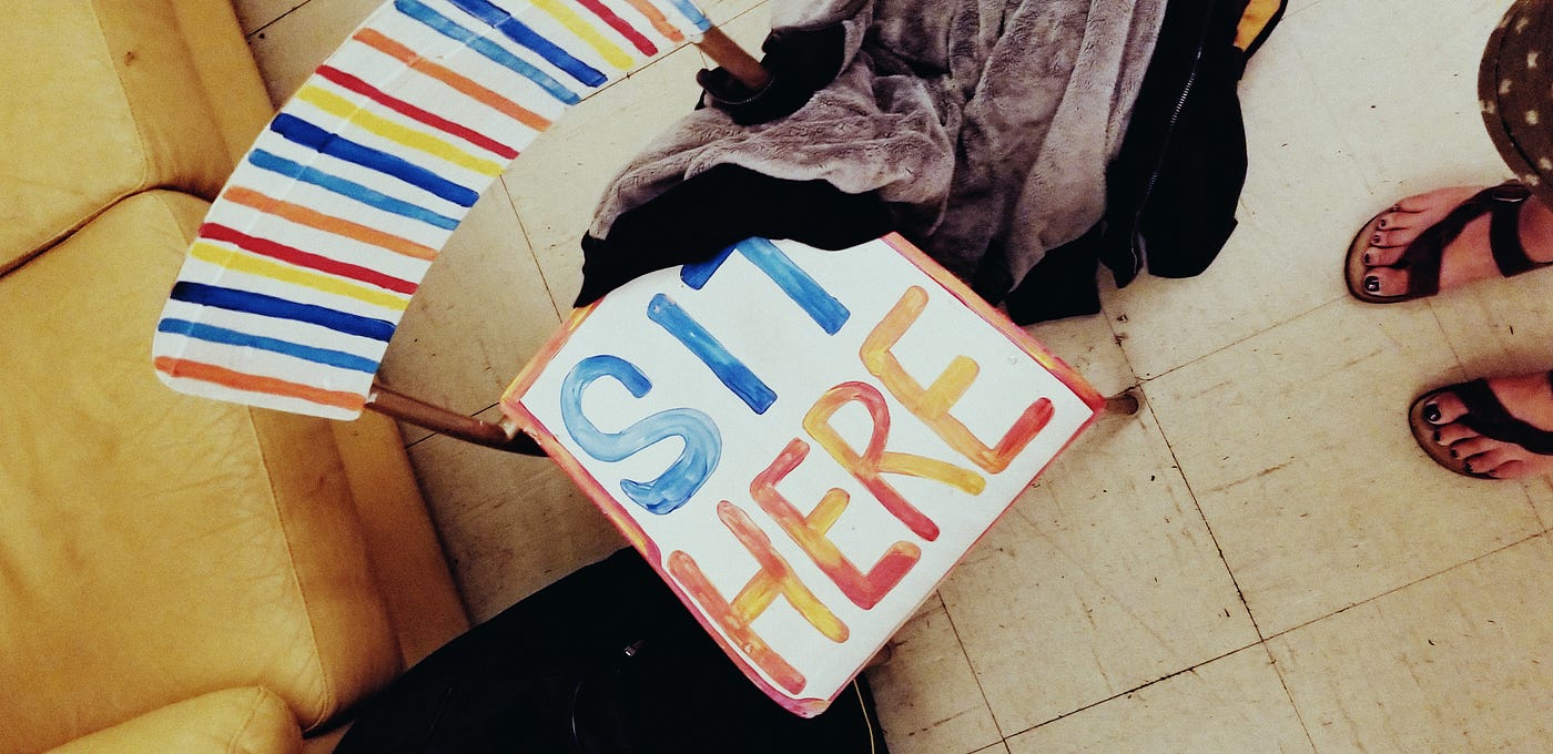 Bird’s eye view of wooden and metal stack chair painted in white, red, orange and blue, with text on seat: “SIT HERE”.