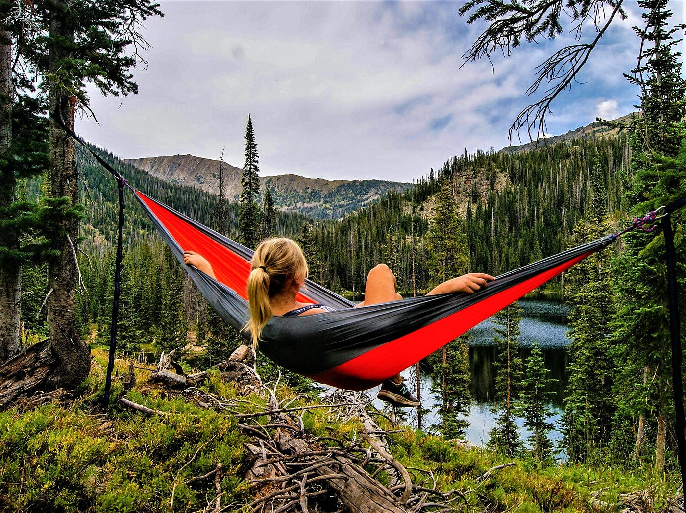 girl with blonde hair in ponytail sitting in red and black hammock looking out over river and forest