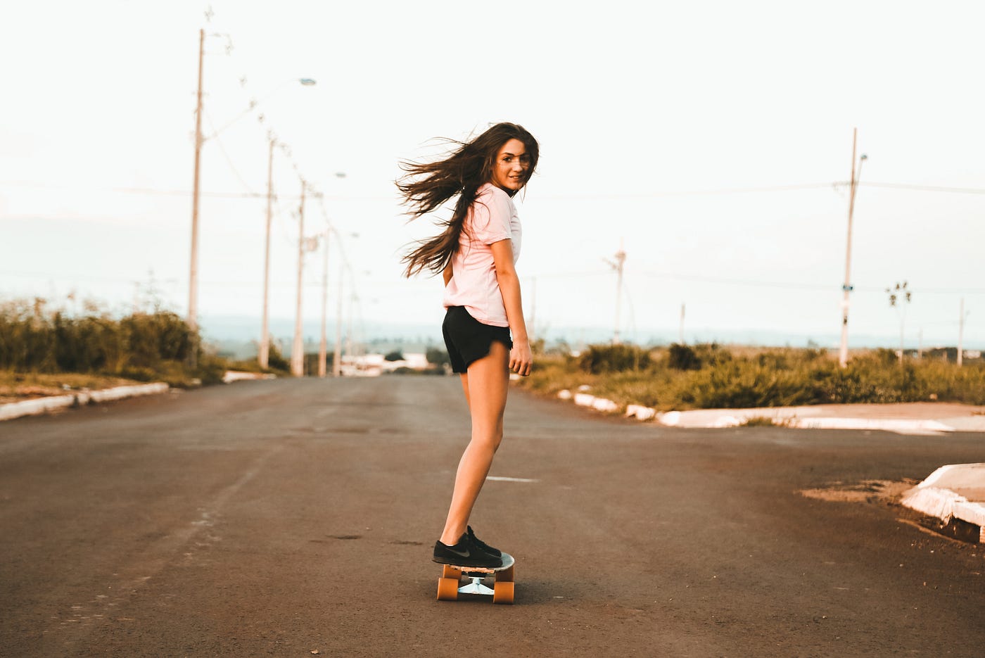 A young woman rides on a skateboard on a wide, empty street, shrubs and curbs decorating the border of the road, a vintage filter over everything.