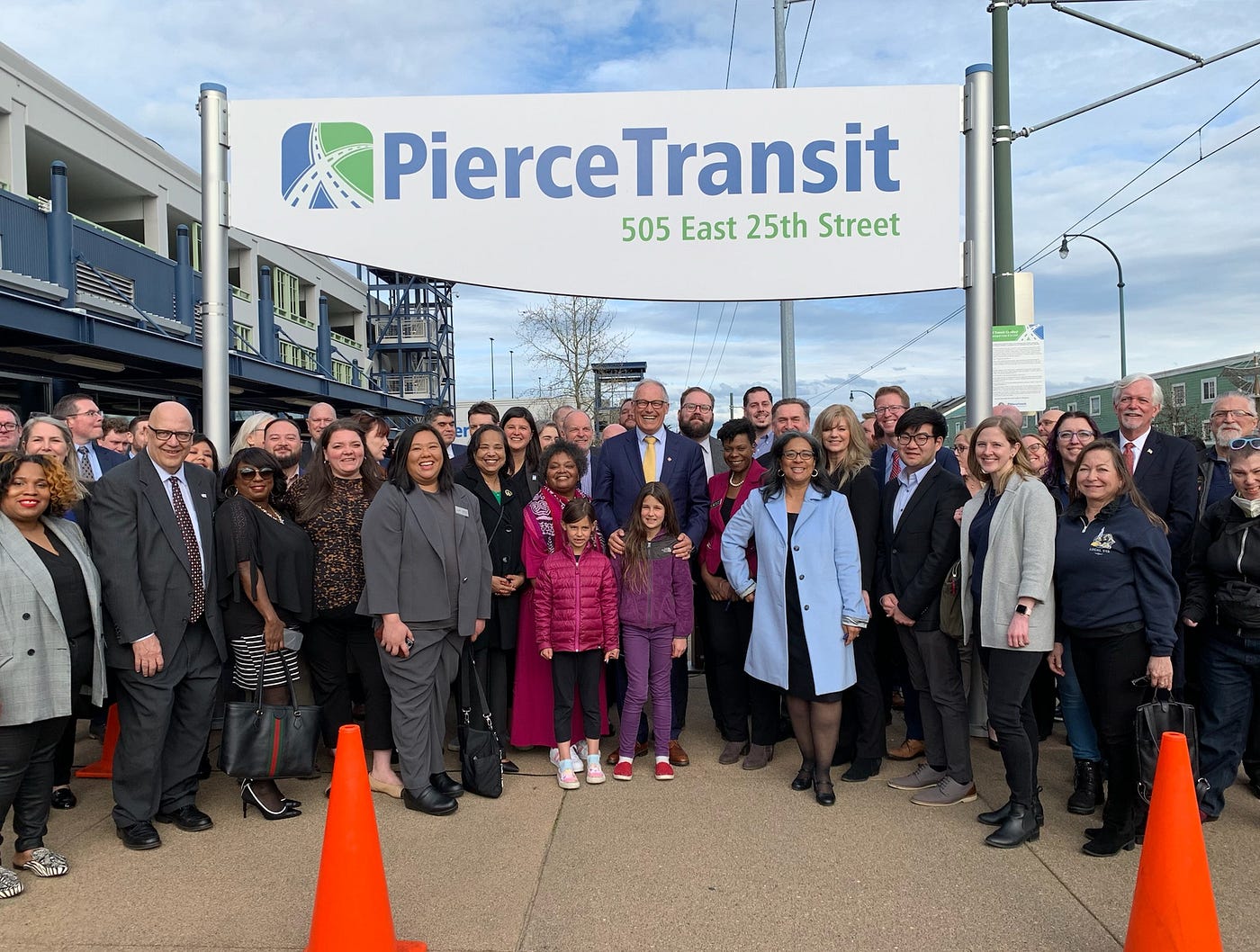 The governor poses with a large crowd of people outdoors. They are smiling at the camera and standing under a large Pierce Transit sign.