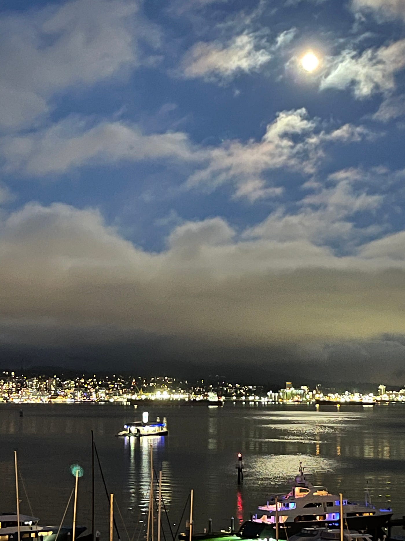 Blue dusk with almost full moon above low clouds, city and boat lights. The moon is reflected zigzagging on the water.