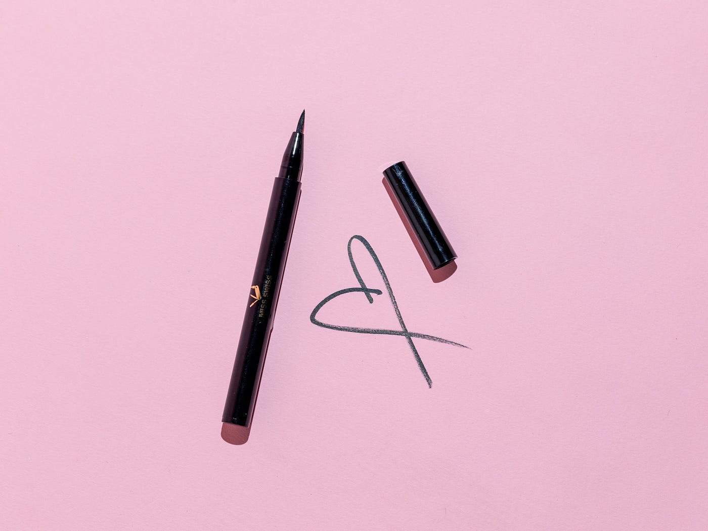 On a pink background, a fountain pen is shown to the left of a drawn heart. To the upper right of the heart is the cap of the fountain pen.