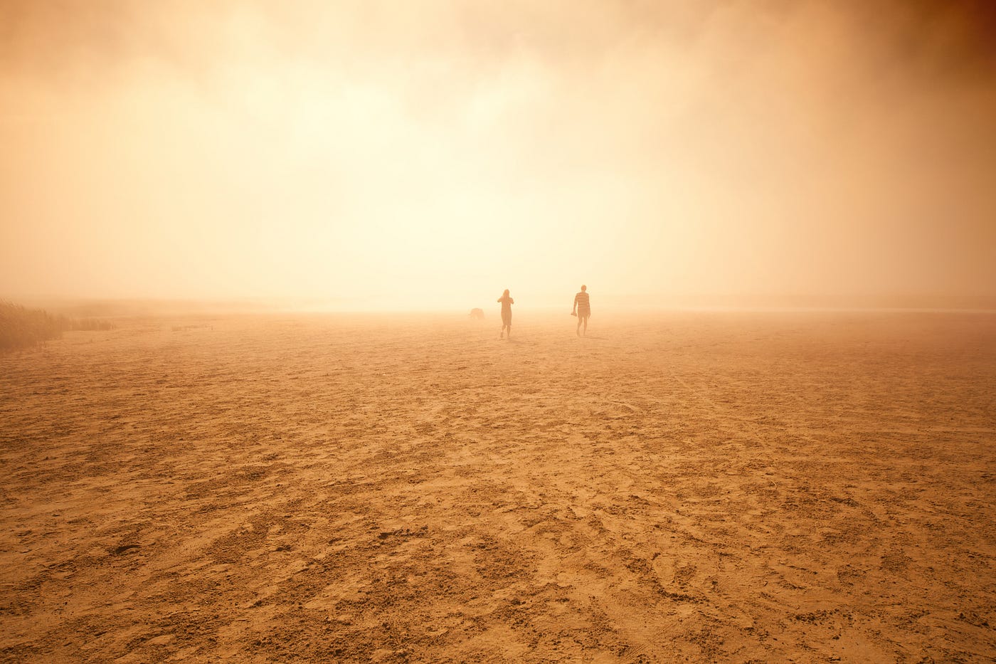 Two people walking in the background of a dessert. The sky is hazy and only the people’s silhouettes are visible.