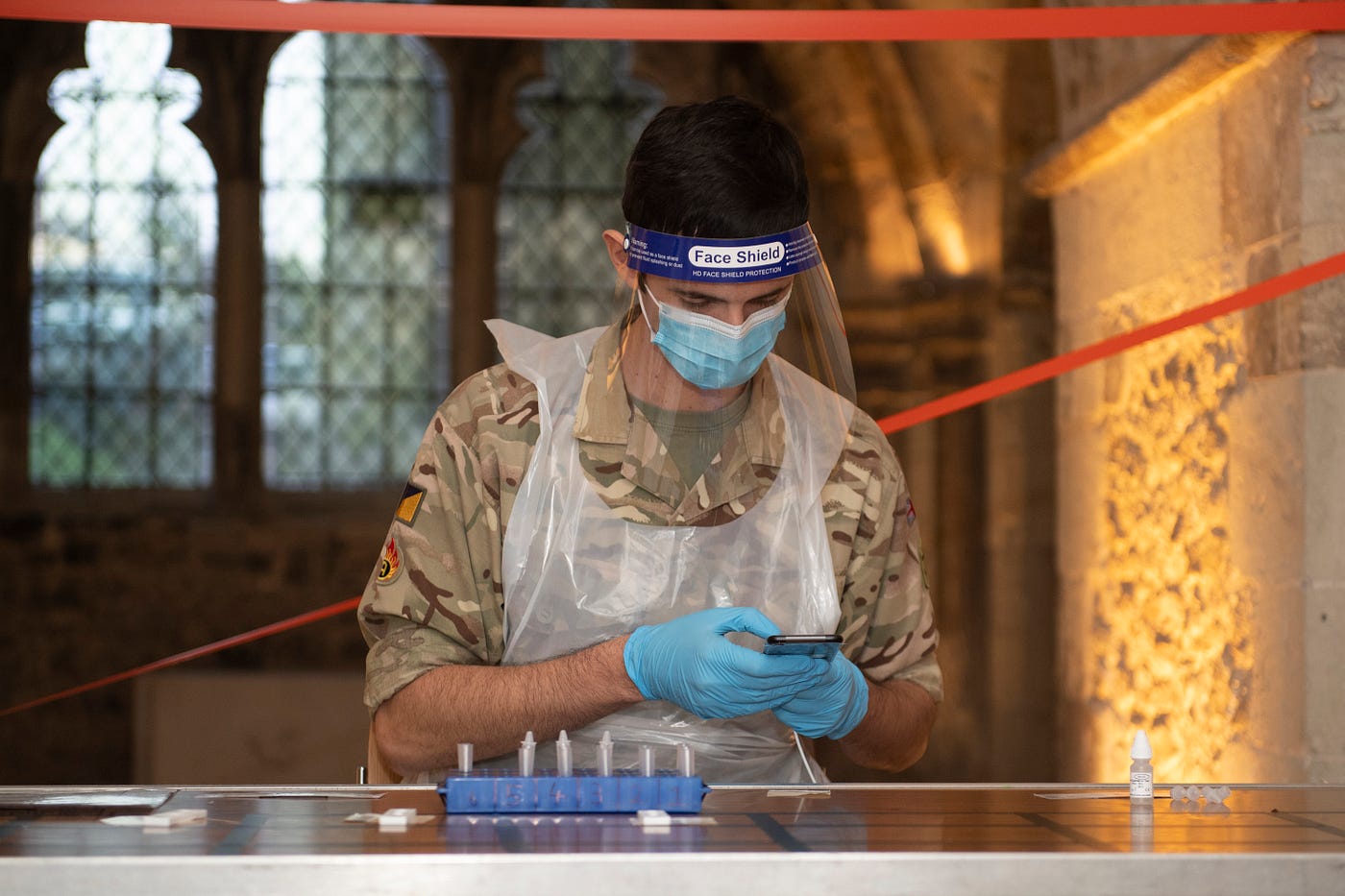 A soldier in a PPE stands in front of a church window processing test tubes, adding information to a mobile device.