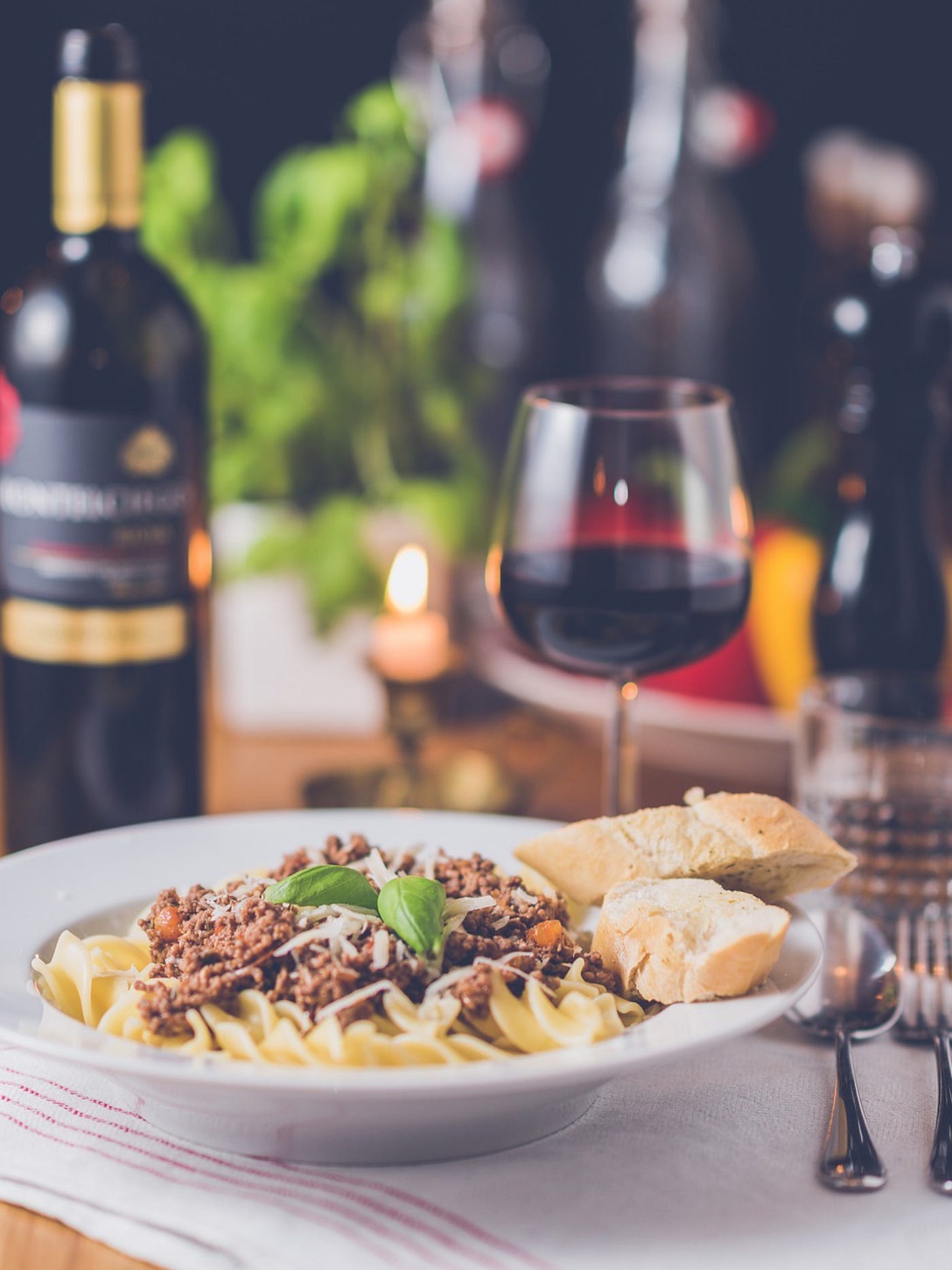 Color photo of a dinner setting, with wine bottle, half-full goblet and a plate of pasta, meat sauce and bread.