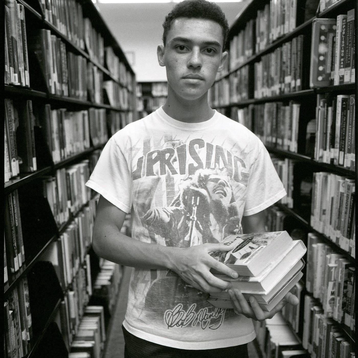 Young man wearing Bob Marley Uprising shirt stands in a library holding books.