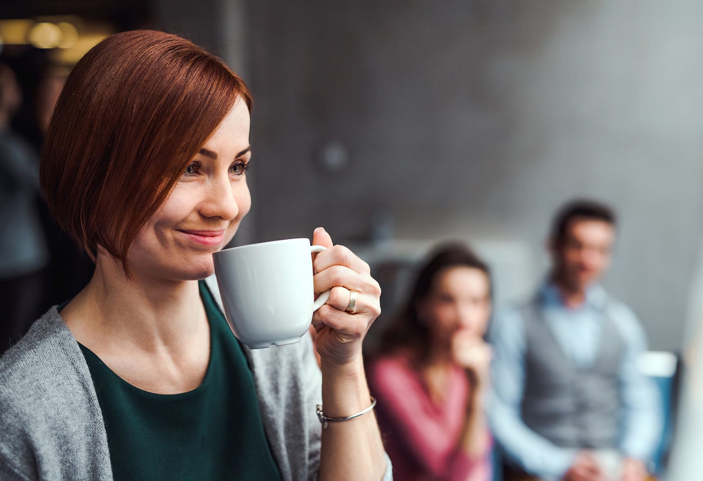 A woman smirks as she holds a coffee mug up to her face in front of 2 coworkers.
