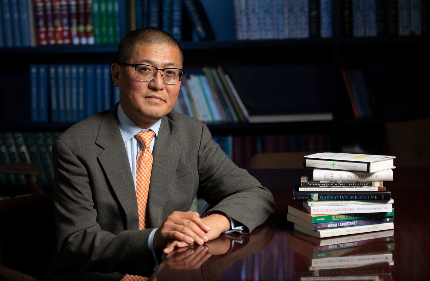 James Kyung-jin Lee rests his hands on the table in front of him, which is stacked with books. Behind him is a book case.