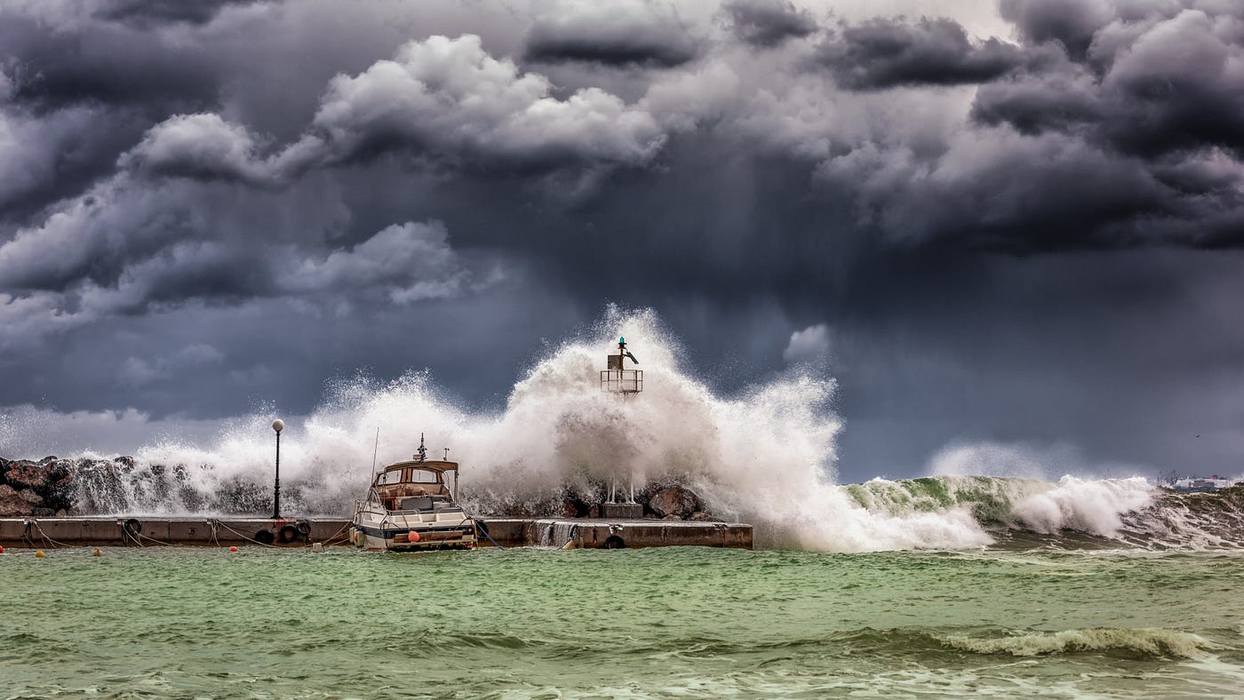 waves crashing against a pier with boats, green water and gray storm sky