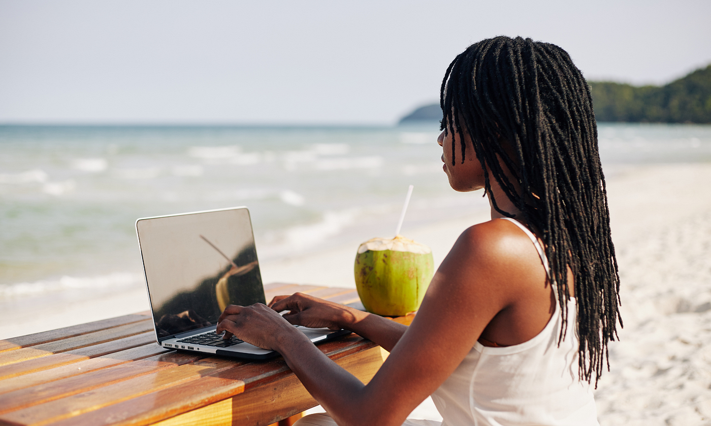 Black woman with long, natural locked hairstyle, typing on a laptop at the beach, drinking from a fresh coconut