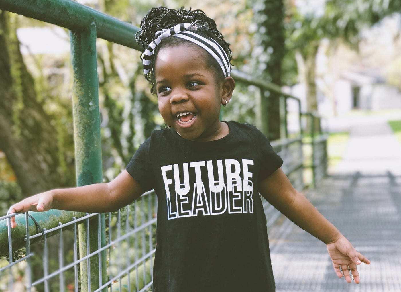 Excited adorable kid, with brown skin and black braids in a striped headband, in a “Future Leader” shirt, on a little bridge