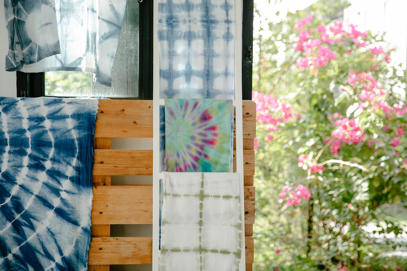 A serene photograph of tie dye hanging on drying racks in blues, pinks, and green colors. The tie dye is hanging inside a house near a window. The window looks out onto a beautiful blurred tree with green leaves and pink flowers. It’s a bright, hopeful, clean aesthetic.