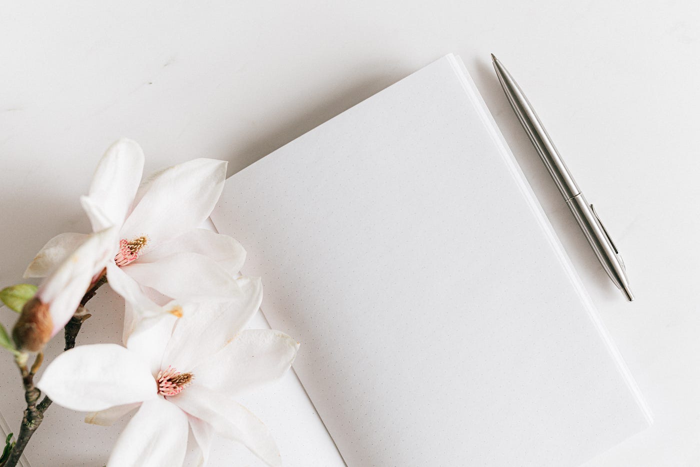 A white journal sits on a smooth white table, a minimalistic silver pen and white and pink flowers next to it.