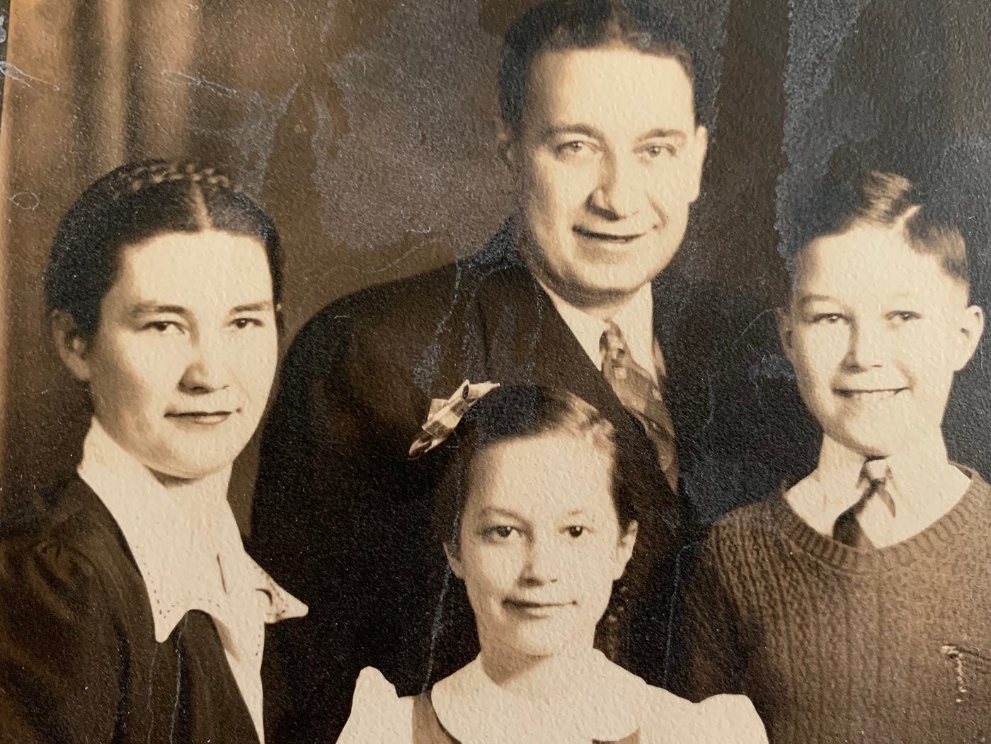 Sepia toned photo from the early 1940s of a white family. My great-grandmother is on the left side with a crown braid, my great-grandfather is in the middle, my great uncle is on the right, and my grandmother is in the bottom center with two pigtail braids and a bow. My great-grandfather and great uncle smile while the females have semi-smiles on their faces.