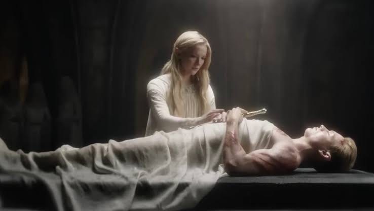 Galadriel da série Anéis de Poder em frente ao corpo deitado de Finrod. Ele segura uma espada dourada com as mãos juntas sobre o peito, que ela toca com delicadeza.