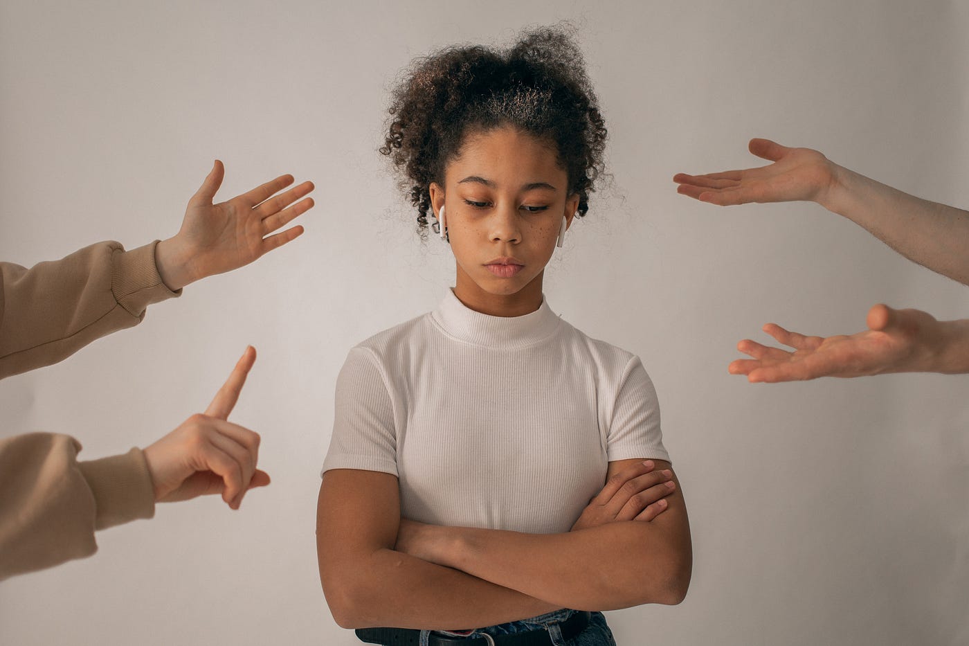 A Black girl stands in the middle of the picture, arms crossed, with headphones in. Around her are white arms and hands pointing towards her, in a seemingly judgy manner.