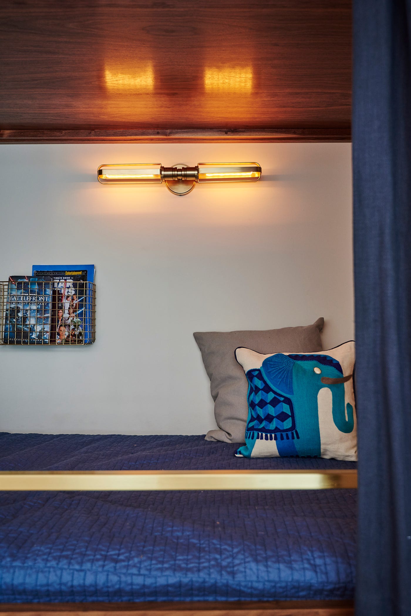 The bottom of a custom walnut bunk bed with a brushed brass bed rail designed by Sarah Barnard, and navy blue linens with black-out lining serving as privacy curtains. An illuminated antique brass wall sconce and matching wall baskets displaying books are mounted above the bed. The navy blue bed is decorated with two pillows — one grey and one decorated with an abstracted elephant in shades of deep blue.