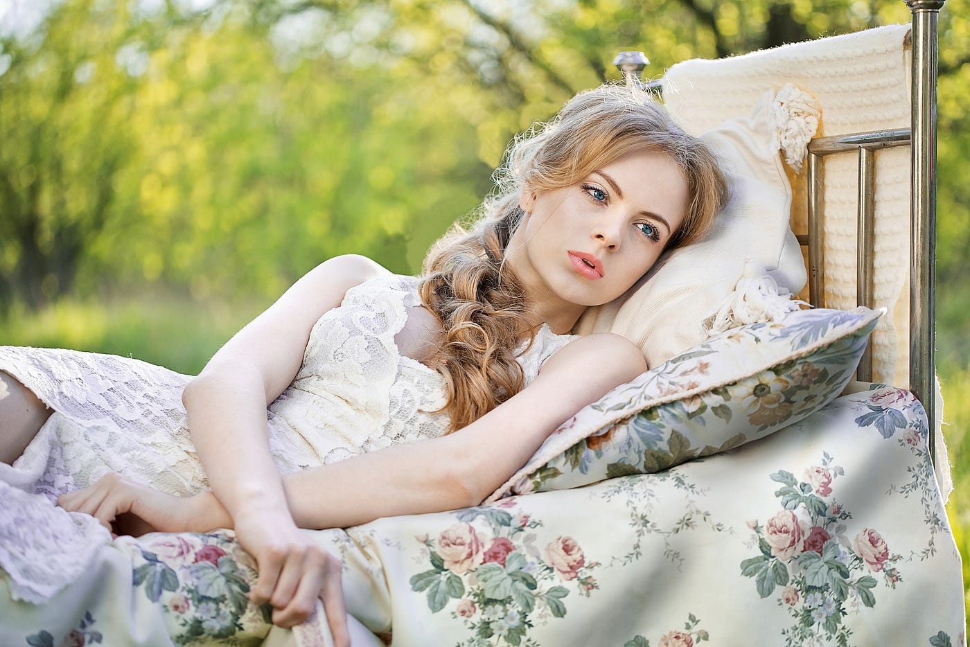 Young woman wearing white dress reclines on a bed with flowered sheets. She looks pensive.