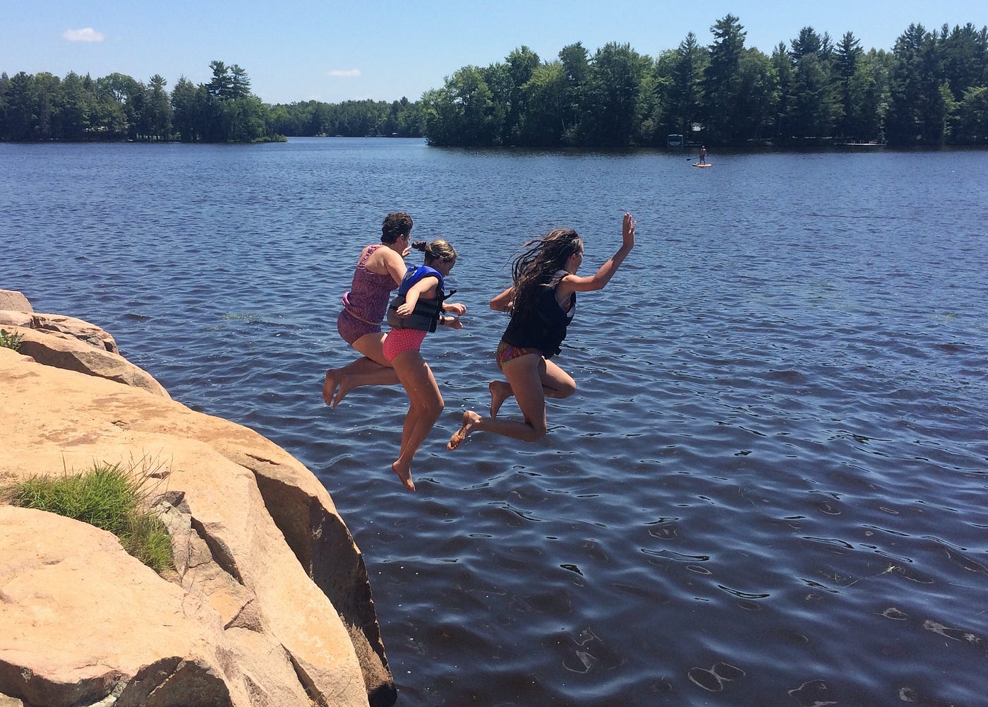 Three people in mid-air, jumping off a large rocky outcropping into a lake.