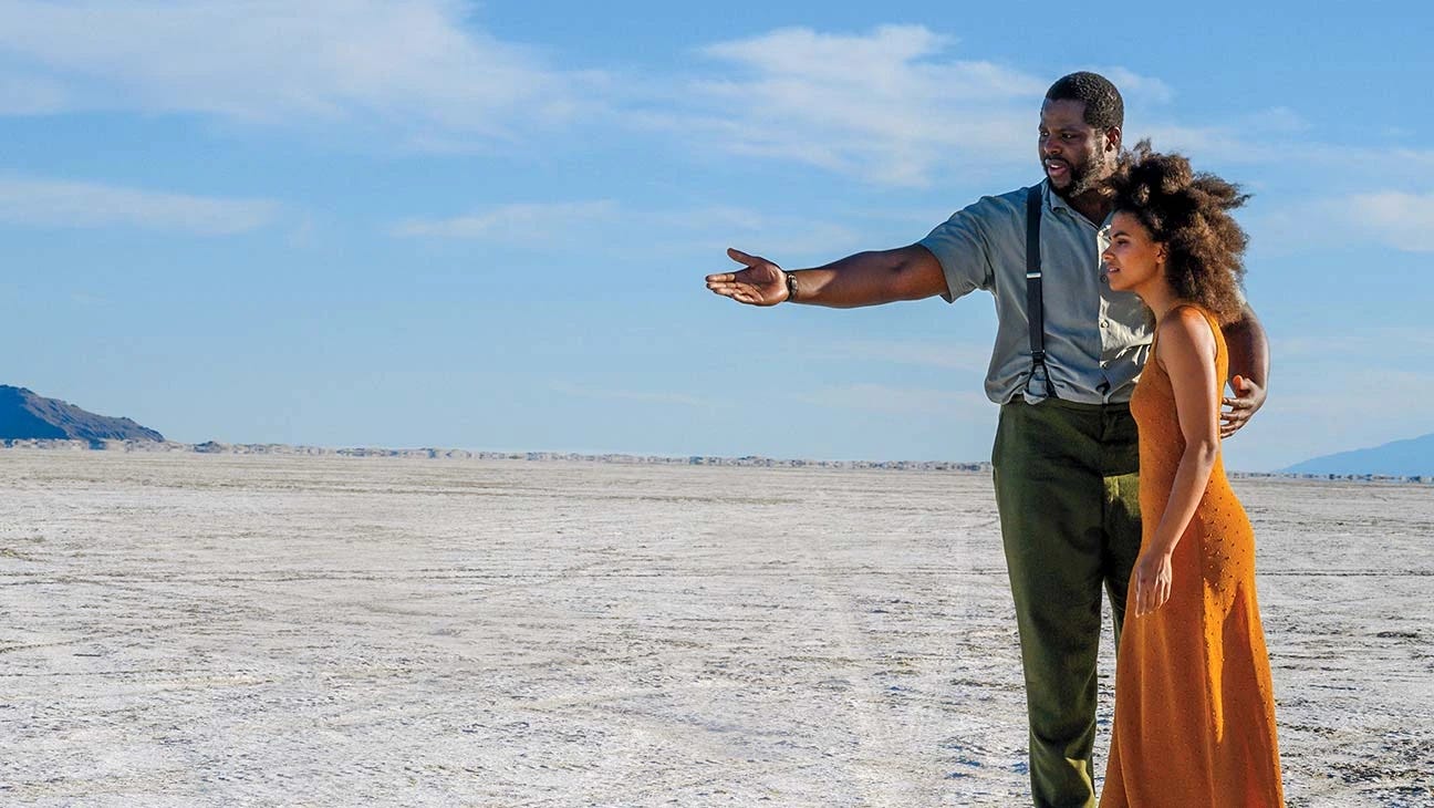 The image features Winston Duke and Zazie Beetz, two African-American actors in the film Nine Days. Duke is dressed in a shirt, pants, and suspenders. Beetz is covered in a long orange dress. The pair stand together on white sand beach with a blue sky backdrop.