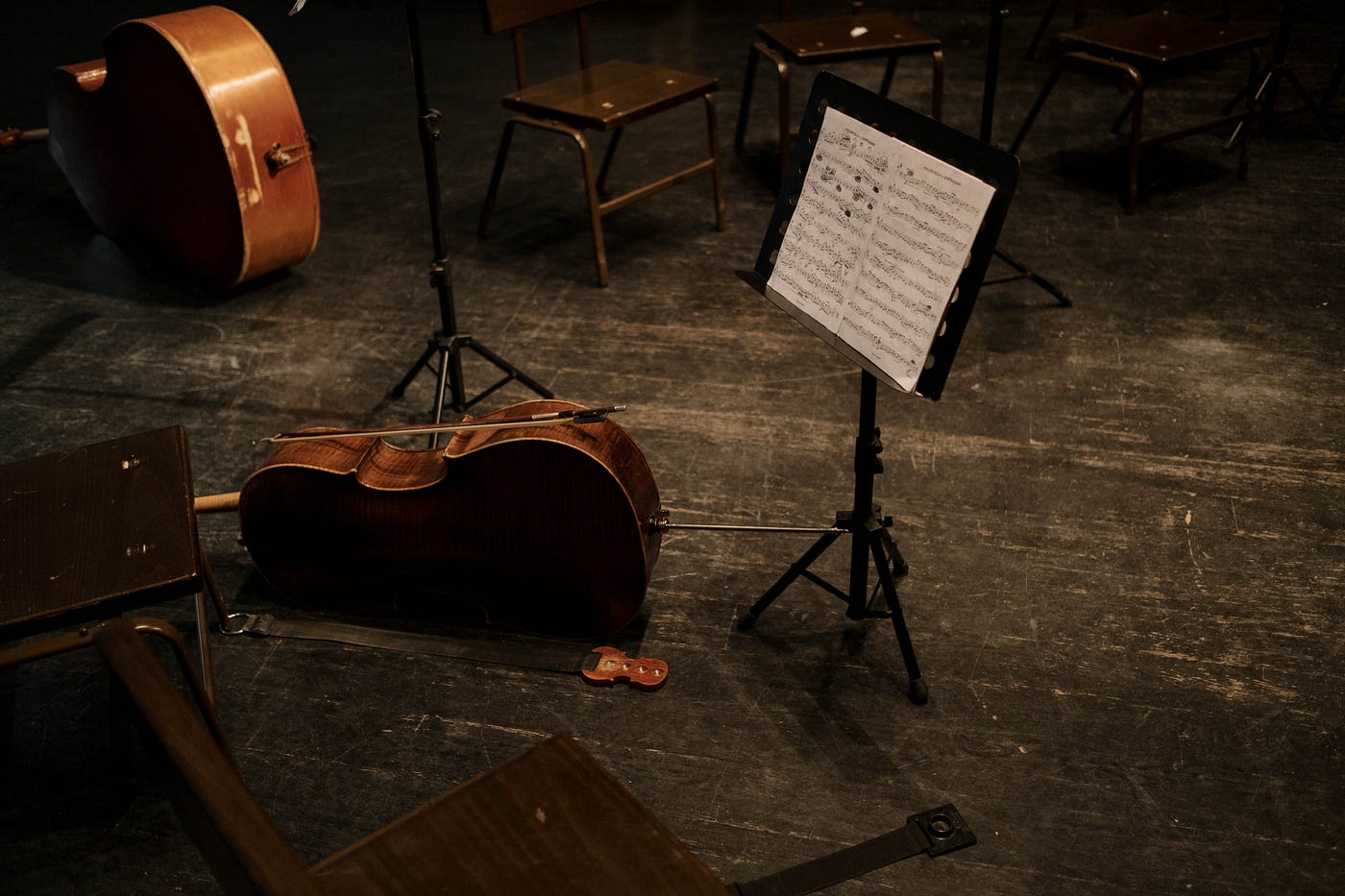 A picture of a music stand on a stage. There is a cello on the ground by the music stand. In the background, there is a bass set down on the ground as well as a few wooden chairs.