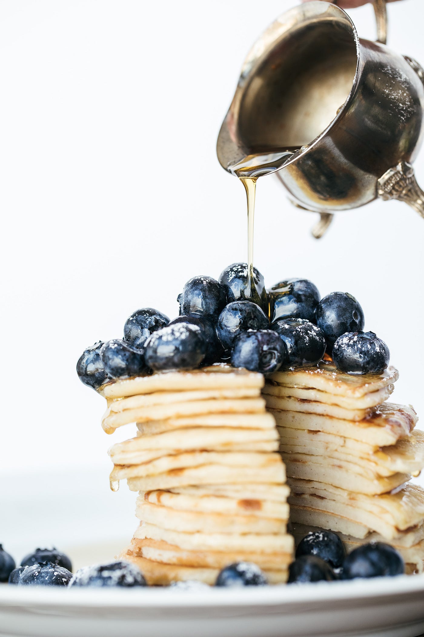 A high stack of pancakes cut in half topped with blueberries and syrup being drizzled from fine china above.
