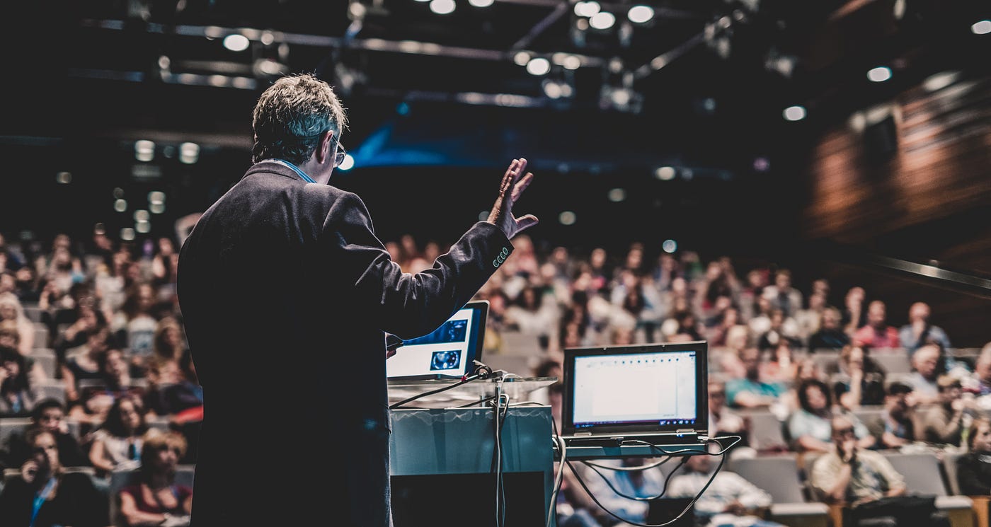 Male speaker in a suit, presenting in front of a room full of people with two computers in front of him.
