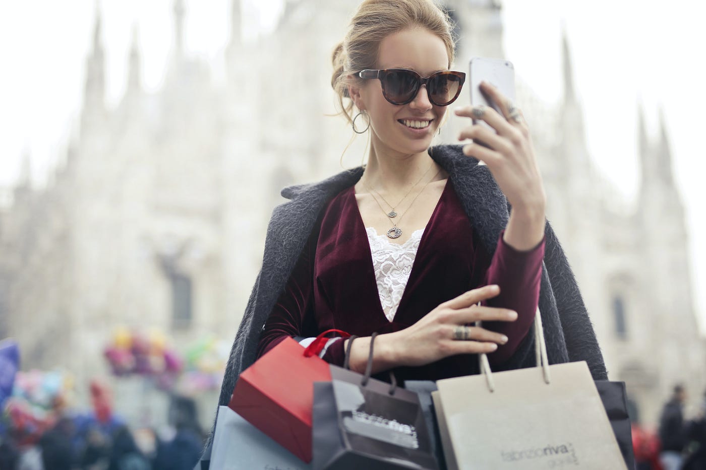 woman wearing sunglasses and holding numerous shopping bags taking selfie.