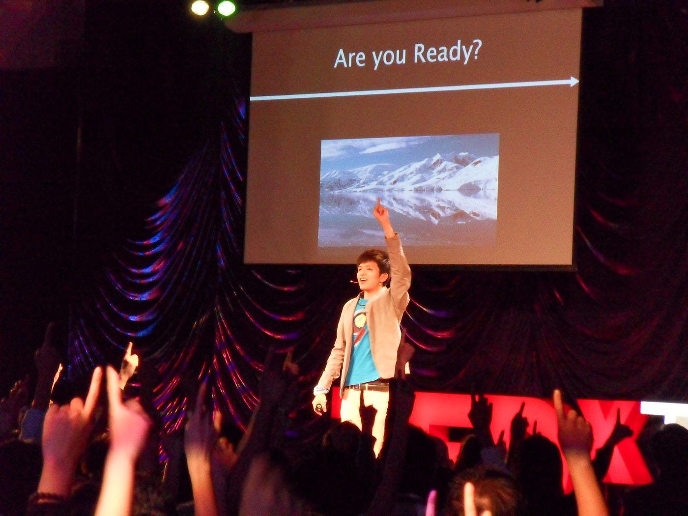 Liang-Yi Chang standing in front of a crowd with one fist raised. A screen behind him shows a photo of glaciers and reads “Are you Ready?” The crowd members’ arms are raised.
