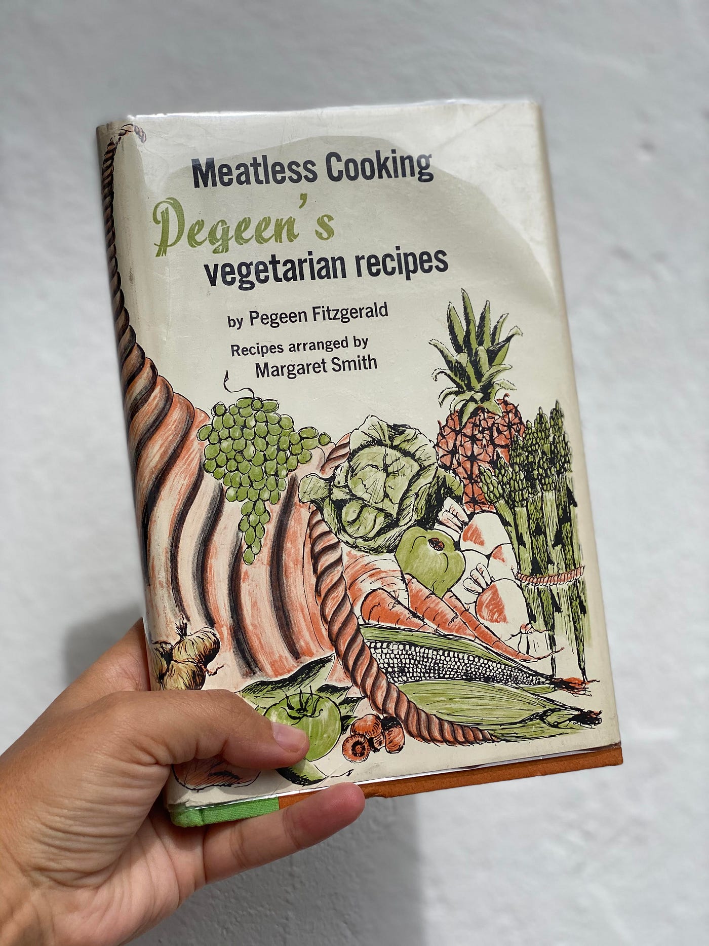 A hand holding up a vintage cookbook with a quaint illustration of fruits and veggies on the front.