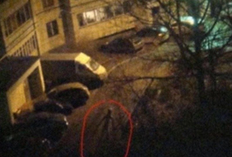 What appears to be a screen capture from surveillance footage. It is night. Cars are parked outside some sort of large building. In the lower center of the image there appears to be a thin “stick figure” shaped humanoid cast in shadow.